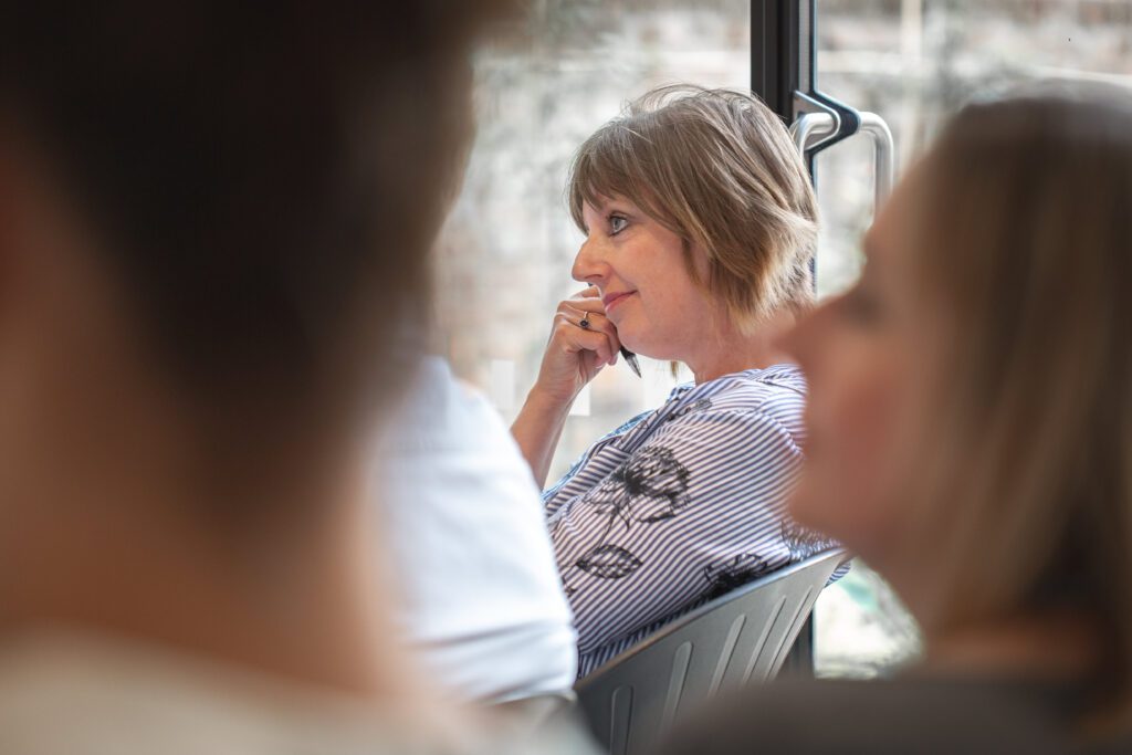 Focus on a woman paying attention to a speaker in a group situation. She is holding a pen and resting her chin on her hand, looking reflective. 